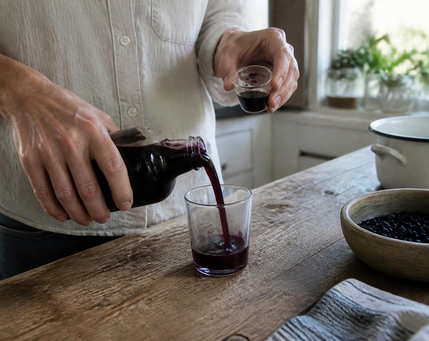 Hands pouring a small 100 ml measure of deep purple aronia juice into a tumbler on a wooden counter