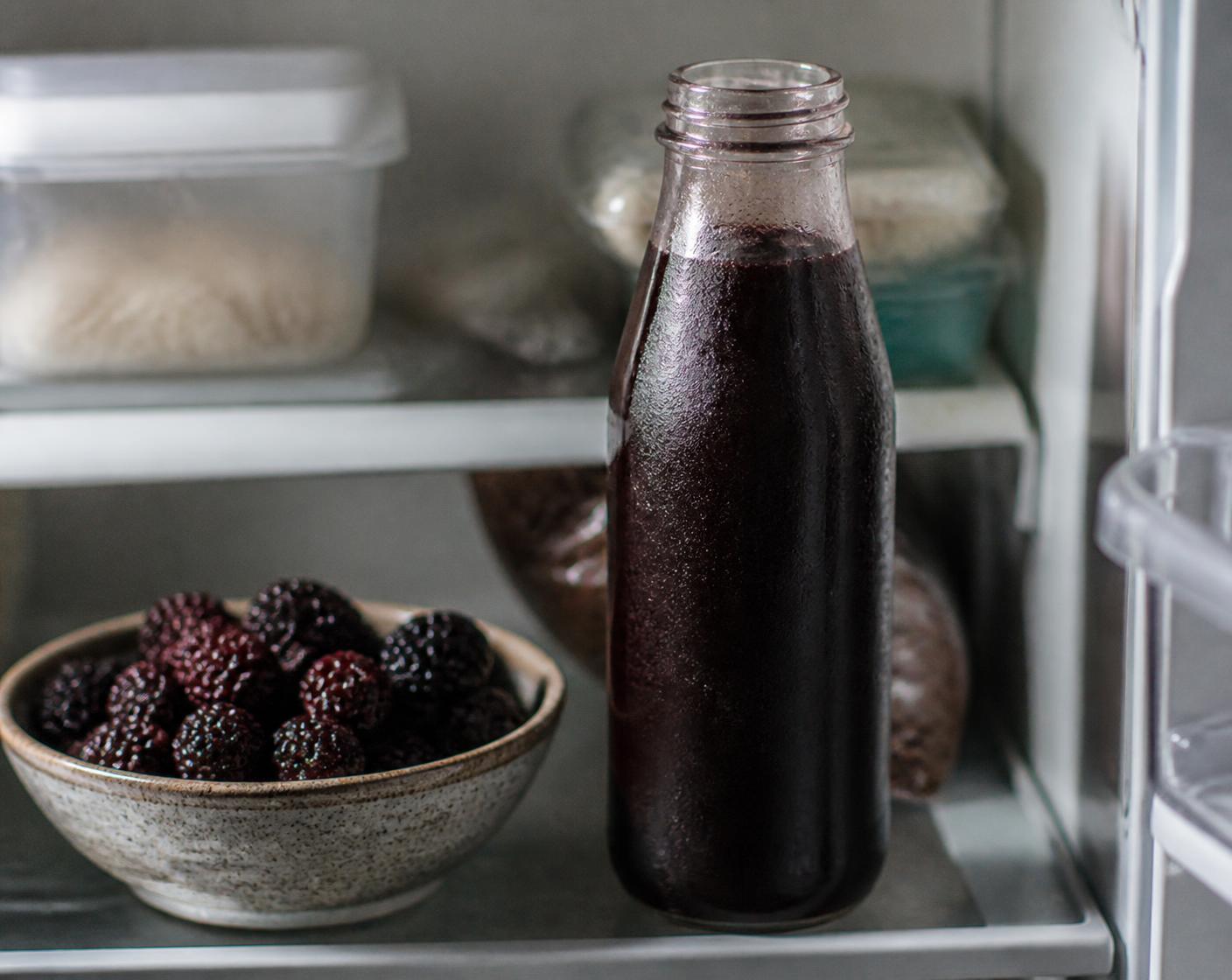 An opened glass bottle of dark aronia juice chilled on a refrigerator shelf beside a bowl of berries